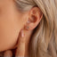 Close-up of an ear wearing a gold crescent moon stud earring with blue stones, the model is pointing to her ear