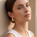 Alegra Botanical Half Hoop Earrings - A close-up lifestyle shot of a woman with her hair pulled back, wearing a textured gold huggie hoop earring, leaning against a beige stucco wall.