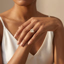 Amore Heart Cluster Stackable Ring Set- A close-up of a woman's hand wearing a gold ring with a white floral cluster setting, styled with a white satin top against a warm beige background.