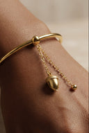 A close-up of a person's wrist wearing a polished gold bangle bracelet with a dangling acorn charm and a small chain accent.
