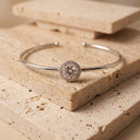 Halo Moissanite Bangle | 1ct VVS Moissanite | 925 Sterling Silver- A polished silver-tone open cuff bracelet with a round halo center stone, displayed on stacked beige stone tiles.