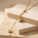 Adora Heart Lariat Necklace- A delicate gold lariat necklace with two polished heart pendants, draped over stacked beige travertine stone blocks.