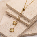Adora Heart Lariat Necklace- A gold lariat-style necklace with two dangling heart charms and a small heart bead at the top, displayed on stacked beige stone tiles.