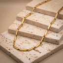 Sculpted Link Necklace – PVD Gold-Plated | Stainless Steel. A gold-toned chain necklace with elongated, polished links draped over stacked rectangular stone tiles on a warm beige surface.