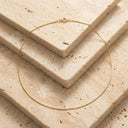 Ivy Barely There Box Chain Necklace. A delicate gold chain necklace with a small lobster clasp, draped elegantly over stacked beige travertine stone tiles.