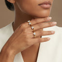 Amore Heart Cluster Stackable Ring Set- A close-up of a model's hand wearing three gold rings, each featuring a small heart-shaped clear gemstone, against a soft beige background.