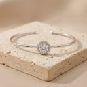 Halo Moissanite Bangle | 1ct VVS Moissanite | 925 Sterling Silver- A polished silver-tone open cuff bracelet with a halo-set round clear gemstone, displayed on a textured beige stone block against a soft beige background.