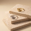 Leaf Open Band Ring – Sterling Silver. Two leaf-shaped rings, one gold and one silver, displayed on stacked beige stone tiles against a warm beige background.