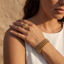 Gold Beaded Bracelet - Stainless SteelA close-up lifestyle shot of a woman wearing a chunky gold ring on one finger and a multi-strand gold beaded bracelet on her wrist, set against a warm sandy background. 