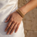 Gold Beaded Bracelet - Stainless Steel -A close-up of a person's hand and wrist wearing a stack of three gold beaded bracelets, resting on a white satin dress against a warm sandy background.