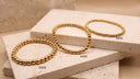 Gold Beaded Bracelet - Stainless Steel- Three gold beaded stretch bracelets displayed on stacked beige stone blocks, labeled 5MM, 4MM, and 3MM to show size variation.