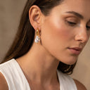 Baroque Pearl Drop Huggie Hoops- A close-up of a woman wearing a gold hoop earring with a white teardrop-shaped pearl featuring small green and black enamel details.