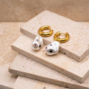 Baroque Pearl Drop Huggie Hoops- A pair of gold huggie hoop earrings with irregular white baroque pearl drops, each adorned with small colourful rhinestones, displayed on stacked beige stone blocks.