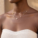 Amara Floral Pearl Station Necklace – Gold or Silver- A close-up of a model wearing a delicate gold necklace with small white pearls and a central round clear stone, paired with a white knit strapless top.
