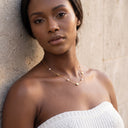 Amara Floral Pearl Station Necklace – Gold or Silver- A woman wearing a delicate gold necklace with small white beads and a tiny gold heart pendant, leaning against a textured beige stone wall.