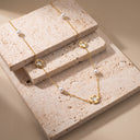 Amara Floral Pearl Station Necklace – Gold or Silver-Two delicate gold chain necklaces with white pearl beads and circular gold pendants are displayed on stacked beige travertine blocks against a warm beige background.