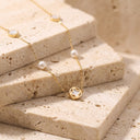 Amara Floral Pearl Station Necklace – Gold or Silver - A delicate gold chain necklace with white pearls and a central gold pendant, styled on stacked beige stone blocks.