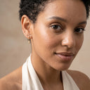 Dainty CZ Drop Chain Stud Earrings - A close-up portrait of a woman with short curly dark hair wearing delicate gold hoop earrings and a small gold drop earring, against a soft beige background.