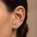 Amelie Rocking Horse Piercing Stud - A close-up of a person's ear showcasing a multi-piece gold ear jewelry set, including a small hoop, a curved barbell with a heart-shaped crystal, a gold ear cuff, and a gold helix band.