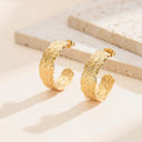 A pair of gold hoop earrings with an embossed leaf pattern, displayed on a light beige surface with a matching stone prop in the background.
