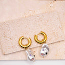 Gold hoop earrings with pearl-like charms on a textured beige surface