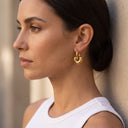 Eliza Convertible Gold Hoops- A close-up side profile of a woman wearing a small gold hoop earring with a heart-shaped cutout, leaning against a textured beige wall.