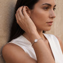 Halo Moissanite Bangle | 1ct VVS Moissanite | 925 Sterling Silver. A woman in a white sleeveless top wears a thin silver cuff bracelet with a round clear gemstone, posed against a textured beige wall.