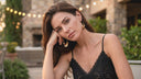 Woman in a black lace top sitting outdoors with a stone building and greenery in the background wearing earrings