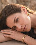Woman wearing gold jewelry including earrings and a bracelet, with a blurred natural background