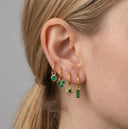 Close-up of an ear wearing gold hoop earrings with green gemstones against a neutral background