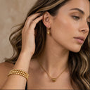 Woman wearing gold jewelry including earrings, necklace, and bracelet against a neutral background