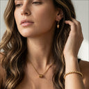 Woman wearing gold jewelry including earrings, necklace, and bracelet against a neutral background