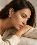Woman wearing Ella and Faith London gold earrings and bracelet, looking thoughtful with a neutral background