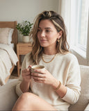 Woman sitting on a couch holding a mug in a cozy room wearing Ella and Faith London Jewellery