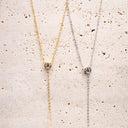 Moissanite Drop Pendant Necklace. Two necklaces, one gold and one silver, on a textured beige surface.