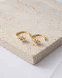 Gold hoop earrings with clear gemstones on a beige stone surface