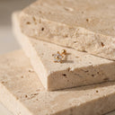 Flora CZ Flower Piercing Stud. A small gold flower-shaped stud earring with clear stones, resting on stacked beige travertine stone slabs.