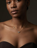 A close-up of a model wearing a delicate gold chain necklace with a small silver-toned flower-like pendant and a tiny pearl accent, paired with a dark strapless top against a dark background.