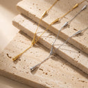 Two delicate chain necklaces with tapered cone pendants, one in yellow gold and one in silver, displayed on stacked beige travertine stone slabs.
