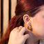 Close-up of a person wearing gold earrings and a ring, with a blurred background, with an ear aid.