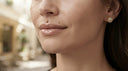 Close-up of a woman's face showing multiple gold body piercings, including a nose stud, lip piercing, and ear cartilage stud with a floral cluster design.