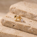 Amelie Rocking Horse Piercing Stud - A gold stud earring shaped like a rocking horse with a clear heart-shaped gemstone, resting on textured beige stone blocks.