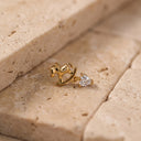 Amelie Rocking Horse Piercing Stud- A gold stud earring featuring a miniature rocking horse and a dangling clear heart-shaped gemstone, resting on beige stone tiles.