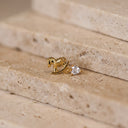Amelie Rocking Horse Piercing Stud- A gold stud earring featuring a small rocking horse and a heart-shaped clear gemstone, resting on beige stone steps.
