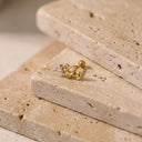 Amelie Rocking Horse Piercing Stud - A gold hoop earring with a small clear gemstone and curved decorative detail, displayed on stacked beige travertine stone slabs.