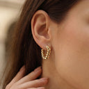 Ayra Twist Hoop Earrings- Gold hoop earring worn by a person with blurred background