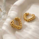 Ayra Twist Hoop Earrings- Two gold-toned twisted hoop earrings resting on a softly draped white fabric surface with a glass object partially visible at the left edge.