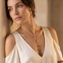 Adora Heart Lariat Necklace- A woman wearing a delicate gold lariat necklace with two small heart charms, styled with a cream cold-shoulder top against a warm beige background.