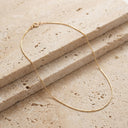 Ivy Barely There Box Chain Necklace. A delicate gold chain necklace with a small clasp, draped elegantly over two stacked rectangular travertine stone slabs.