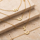 Gold Initial Bracelet -four gold initial necklaces with lowercase-style letters 's', 'c', and 'j' draped over stacked beige stone blocks on a textured beige surface.